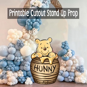 Puede incluir: Un accesorio de pie recortado e imprimible con Winnie the Pooh en un tarro de miel con la palabra "HUNNY". El accesorio está rodeado por un arco de globos en tonos azules y blancos, con acentos florales. El texto "Printable Cutout Stand Up Prop" se muestra en la parte superior.