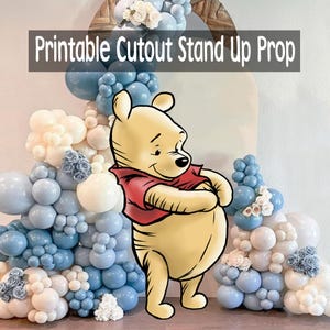 Puede incluir: Un accesorio recortable imprimible con Winnie the Pooh, con una camisa roja, de pie frente a un arco de globos en tonos azules y blancos. El texto "Printable Cutout Stand Up Prop" se muestra en la parte superior.