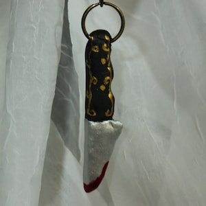 May include: A black and gold knife keychain with a silver blade and a red tip. The keychain is hanging from a gold chain.