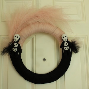 May include: A black wreath with pink pampas grass and two skeleton figures with white faces. The wreath is hanging on a white door.