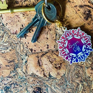 Könnte beinhalten: Ein Schlüsselbund mit einem dekorativen Schlüsselanhänger. Der Anhänger hat ein kreisförmiges Design in Rot-, Lila- und Blautönen mit dem Schriftzug "Bugger Off". Die Schlüssel liegen auf einer braunen, strukturierten Oberfläche.