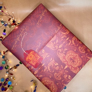 Puede incluir: Un regalo envuelto en papel burdeos con un diseño de cachemira dorado. El regalo tiene una etiqueta a juego. El texto "Enjoy the gift you twat" está impreso en el papel de regalo. Una guirnalda de luces de colores está en el fondo.