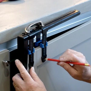 Puede incluir: Una herramienta negra y azul con un mango plateado se utiliza para marcar una puerta de armario con un lápiz rojo. La herramienta se utiliza para asegurar que las bisagras se coloquen en la posición correcta.