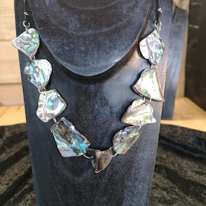 May include: A necklace made of iridescent abalone shell pieces, each with a unique shape and pattern, connected by silver rings. The necklace is displayed on a black mannequin bust.