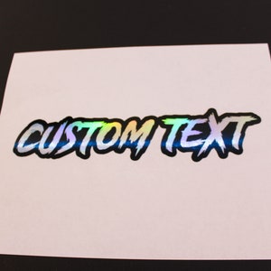 May include: A holographic sticker with the text "CUSTOM TEXT" in a black and white stylized font. The sticker has a rainbow iridescent finish.