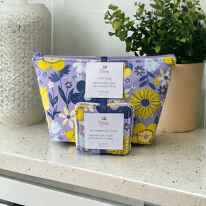 Puede incluir: Una bolsa de tocador morada con un estampado floral y flores amarillas. La bolsa está hecha de 100% algodón con un forro impermeable. Viene acompañada de dos toallitas faciales reutilizables hechas de 100% algodón y bambú.