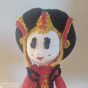 Puede incluir: Una muñeca de crochet que lleva un vestido rojo con detalles dorados y negros. La muñeca tiene el pelo negro y una cara blanca con ojos negros y una boca roja.