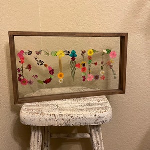 May include: A framed piece of art featuring the word "SUMMER" spelled out using pressed flowers in various colors, including pink, yellow, blue, and green. The frame is brown wood and the background is white.