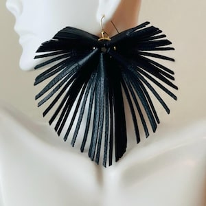 May include: A large, fan-shaped earring constructed from numerous thin, black leather strips radiating from a central gold-toned metal ring. The design features a textured surface on the leather fringe, creating a wide, layered silhouette that hangs from a simple gold-toned hook wire.