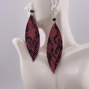 Peut inclure: Paire de boucles d'oreilles pendantes en forme de feuille. Elles pr&eacute;sentent un motif peau de serpent rose et noir. Chaque boucle d'oreille est orn&eacute;e d'un crochet dor&eacute; et d'une petite perle bleue.