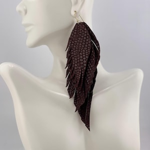 May include: Burgundy leather feather earrings with a textured, scale-like pattern. The earrings have a gold-colored hook and are displayed on a white mannequin. The earrings are long and dangle.