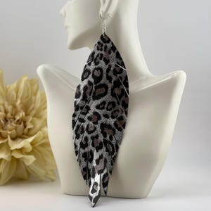 May include: Large, silver and black leopard print feather earrings. The earrings are displayed on a white mannequin bust. The earrings have a silver hook and are made of a material that resembles leather.