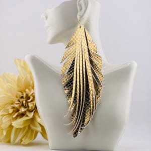May include: Gold-colored feather earrings with a textured, snake-skin pattern. The earrings are displayed on a white bust, with a yellow flower in the background. The earrings have a layered design with white accents.