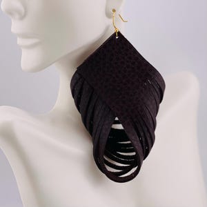May include: A dark brown, diamond-shaped earring featuring a textured surface and multiple layered fringe strips that loop downward to create a hollow, rounded center. The earring is attached to a gold-toned metal hook for ear placement.