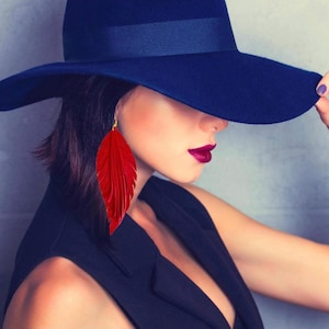 May include: A long, vibrant red feather earring with a gold-toned hook attachment. The earring features a layered, leaf-like structure with fine, detailed textures that mimic the appearance of a natural feather, hanging from a gold metal loop.