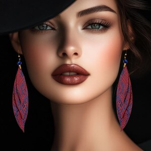 Peut inclure: Portrait d'une personne portant des boucles d'oreilles en forme de plumes, &agrave; motifs de peau de serpent rouge et violet, avec des accents bleus. La personne a les cheveux fonc&eacute;s, un chapeau noir et du rouge &agrave; l&egrave;vres fonc&eacute;.