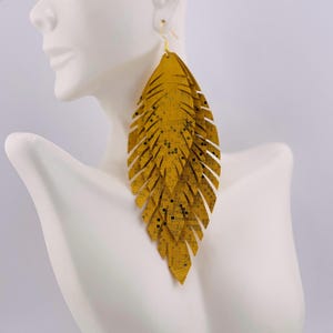 May include: Mustard yellow feather-shaped earrings with gold-colored hooks. The earrings have a layered design with a textured surface and small black square accents. The earrings are displayed on a white mannequin.