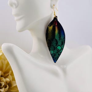 May include: A single, large, leaf-shaped earring with a gold hook. The earring displays a gradient of colors, including green, blue, yellow, and red, with a textured, scale-like pattern. The earring is displayed on a white mannequin.