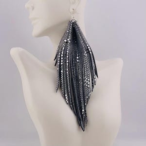 May include: Large, dangling earrings with a dark gray, fringed design. The earrings have a textured pattern with silver accents and are displayed on a mannequin. The earrings are designed to be worn with a hook.
