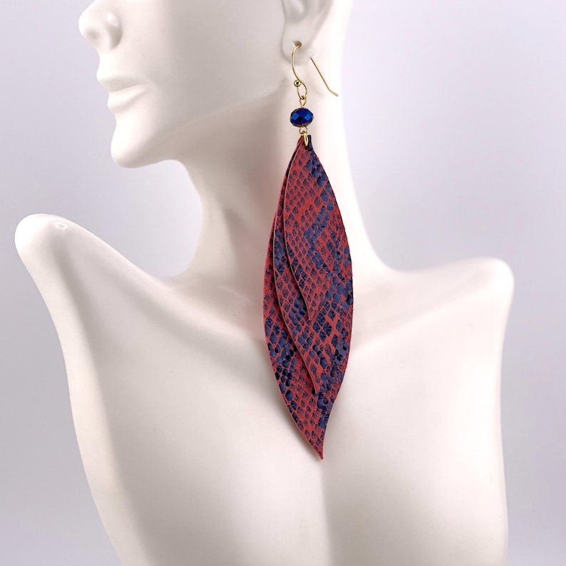 Peut inclure: Longues boucles d'oreilles pendantes avec un motif peau de serpent rouge et bleu marine. Les boucles d'oreilles sont en forme de feuille et sont suspendues &agrave; des crochets dor&eacute;s. Une petite perle bleu fonc&eacute; orne le haut de chaque boucle d'oreille.