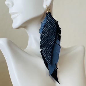 May include: Dark blue leather feather earrings with gold-colored ear wires. The earrings feature layers of textured and smooth leather, creating a layered feather design. The earrings are shown on a white mannequin.