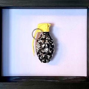 以下が含まれることがあります： 装飾的な手榴弾を特徴とする額入りのアート作品。手榴弾は、白い花柄の黒いベースと黄色のトップを備えています。アートワークは、黒いフレーム内の白い背景に対して設定されています。