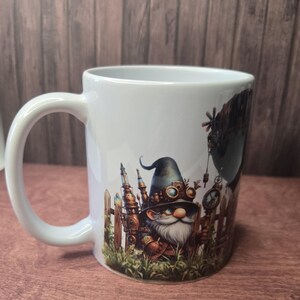 Könnte beinhalten: Ein weißer Keramikbecher mit einer farbenfrohen Grafik eines bärtigen Gnoms, der einen dunklen Hut mit Schutzbrille trägt und in einer Grasfläche vor einem Holzzaun und mechanischen Strukturen steht, darüber ein großes dunkles Luftschiff.