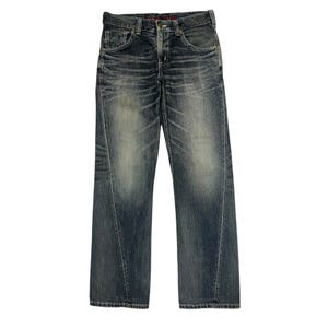 Puede incluir: Vaqueros de mezclilla azul oscuro con un efecto de lavado desgastado en los muslos y las rodillas. El pantalón incluye bragueta de botones, bolsillos delanteros y detalles de costuras verticales a lo largo de las piernas, con un logo rojo.
