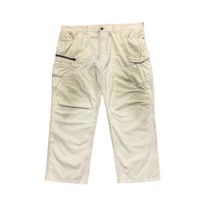 Puede incluir: Pantalones cargo color crema con múltiples bolsillos y cierre de cremallera en la pierna izquierda. Los pantalones tienen un corte holgado y cierre de botón y cremallera en la cintura. El texto "WORLD STANDARD EQUIPMENT U.S.A." está impreso en el bolsillo izquierdo.