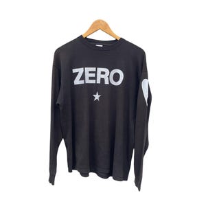May include: A black long-sleeve shirt featuring the word ZERO printed in white across the chest with a white star graphic positioned directly below the text. The left sleeve displays a white heart-shaped graphic near the shoulder area.