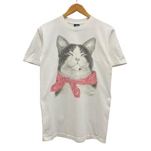 May include: White t-shirt featuring a smiling cat with a black and gray face, wearing a pink bandana. The graphic is a detailed illustration. The shirt is hanging on a wooden hanger.