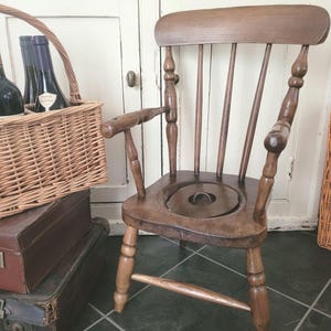 May include: A wooden potty chair with a brown finish. The chair has a simple design with a rounded seat and a small hole in the centre. The chair is sitting on a tiled floor.