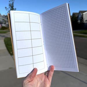 May include: An open notebook with a brown cover featuring white pages. The left page displays a grid layout divided into eight rectangular sections, while the right page features a full-page grid pattern with thin black lines on a white background.