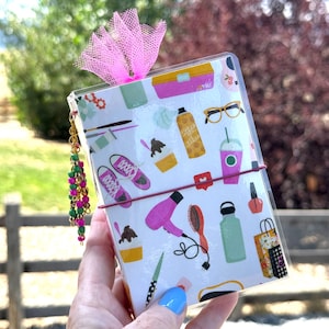 May include: A rectangular notebook with a clear plastic cover featuring a white background with colorful illustrations of sneakers, a hair dryer, a hairbrush, a water bottle, glasses, and shopping bags. A pink tulle bow and a beaded charm are attached.
