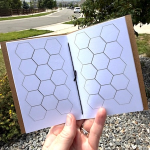 May include: A small notebook with a brown cover and white interior pages featuring a black ink grid of repeating hexagons. The pages are held together by a thin black elastic cord running vertically through the center fold of the booklet.