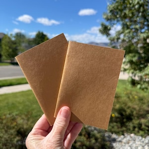 May include: Two rectangular, folded booklets made of plain, light brown kraft paper. Each booklet features a simple, minimalist design with a smooth, matte texture and clean, sharp edges, presented as a pair of identical stationery items.