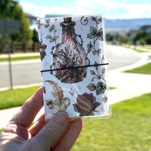 May include: A rectangular notebook encased in a clear plastic cover, secured by a thin black elastic band. The cover features a white background with a repeating illustration of a glass potion bottle, dark roses, and leafy vines with small stars.