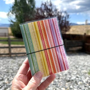 May include: A small rectangular notebook featuring a vertical striped pattern in shades of green, yellow, orange, pink, and red. The cover is protected by a clear plastic sleeve and held closed by a thin black elastic band wrapped around the center.