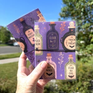 May include: Two rectangular cards featuring a purple background with a repeating pattern of potion bottles labeled Deadly Nightshade, Spider Venom, Elixir No. 13, Trick or Treat, Bat Wings, and Fear No Evil, held together by a thin black elastic band.