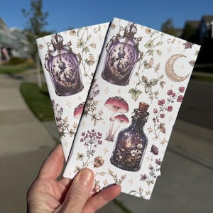 May include: Two rectangular notebooks with a white background featuring a repeating pattern of purple glass potion bottles, pink mushrooms, delicate floral vines, and a crescent moon. The illustrations are detailed with fine lines and soft colors.