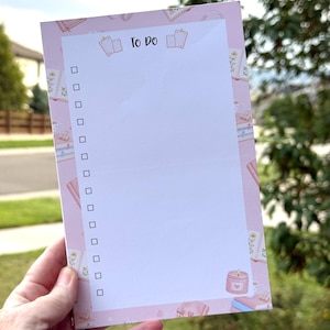 May include: A rectangular notepad with a light pink border featuring small illustrations of books, flowers, and candles. The white center section is headed with the text To Do and includes a vertical column of fifteen empty checkboxes for tasks.