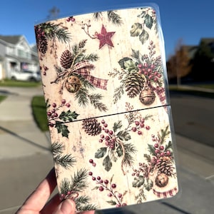 May include: A rectangular notebook with a clear plastic cover featuring a festive print of pine branches, pinecones, red berries, jingle bells, and a red star on a light wood-grain background, secured by a thin black elastic band.
