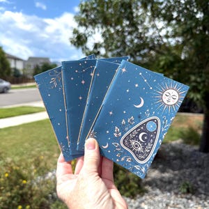 May include: A set of four small blue notebooks featuring a celestial design with a sun, moon, stars, and a planchette graphic in white and gold ink. The notebooks are bound with visible stitching along the spine and have a matte finish.