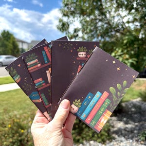 May include: A set of small, dark purple notebooks featuring a printed illustration of a bookshelf with colorful books, potted plants, and gold stars. The notebooks are bound with white thread along the spine and have a matte finish.