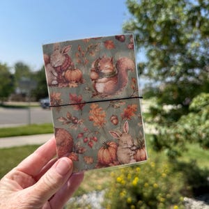 May include: A rectangular notebook encased in a clear plastic sleeve, featuring a repeating pattern of rabbits, squirrels, pumpkins, acorns, and autumn leaves in shades of orange, brown, and green, secured by a thin black elastic band.