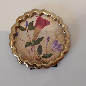 May include: A round coaster with a scalloped gold-toned metal edge. The center features a pressed floral design on a beige background, showcasing a prominent pink flower, smaller purple blossoms, and green leaves preserved under a clear surface.