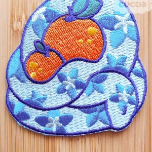 May include: An embroidered iron-on patch featuring a blue and white floral design with an orange fruit in the centre. The patch is perfect for adding a touch of whimsy to clothing, bags, or other fabric items.