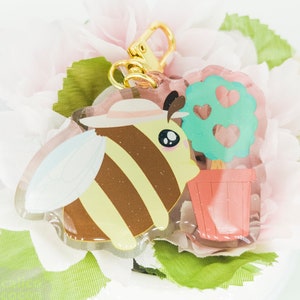 May include: A yellow and brown bee with a white hat and a small tree with pink hearts in a pink pot. The bee and tree are attached to a gold key chain.