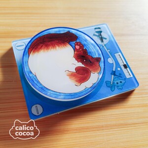 May include: A blue and white circular vinyl record with a white cat curled up on it. The cat has brown and white fur and is sleeping. The record is on a blue and white turntable with a small fish design.