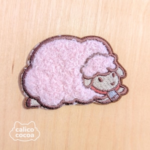 Pode incluir: Patch de aplique de ovelha bordada de cor rosa com costuras marrons. A ovelha está deitada com a cabeça virada para o lado.
